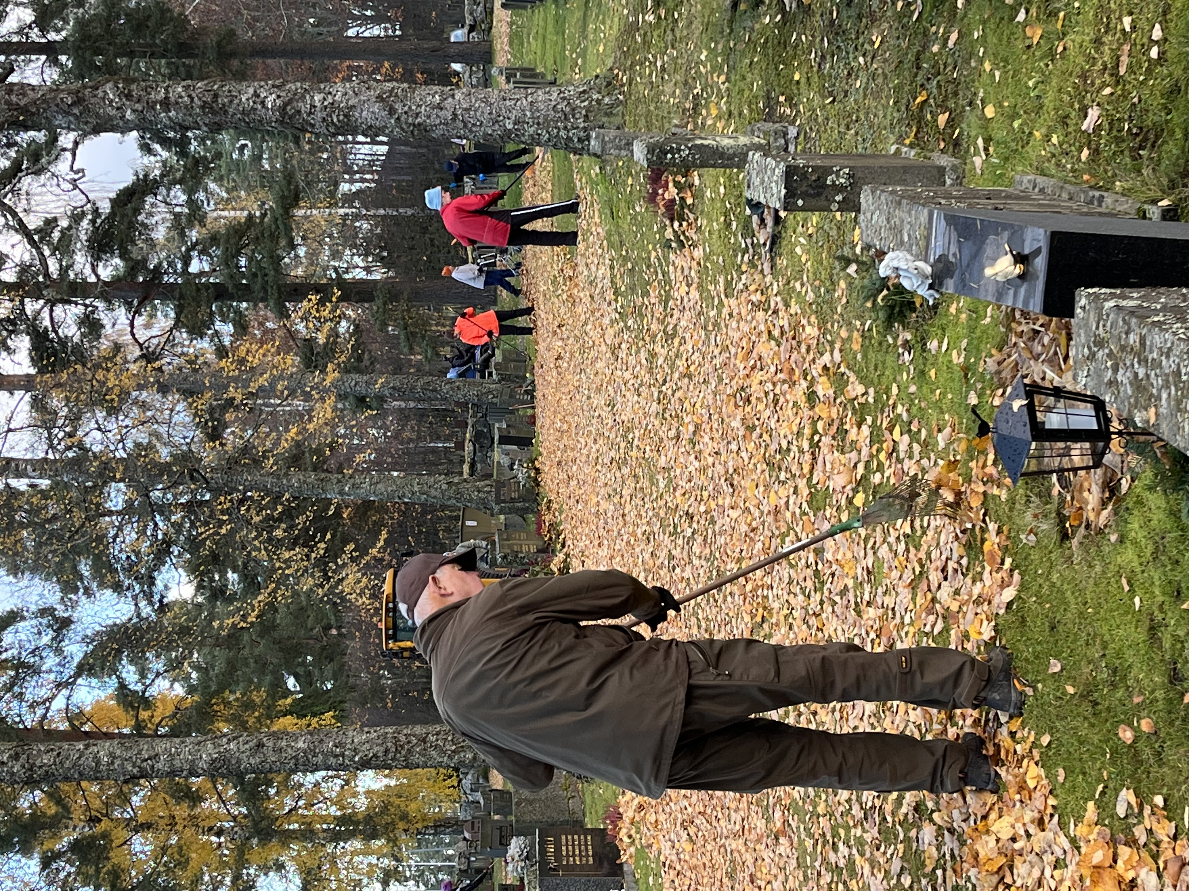 hautausmaalla syksyisiä lehtiä ja henkilöitä haravoimassa